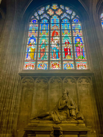 The image features a large stained glass window depicting various religious figures, with vibrant colors and intricate designs. Below the window is a prominent stone statue of a seated figure in contemplative pose, set against a textured stone wall.