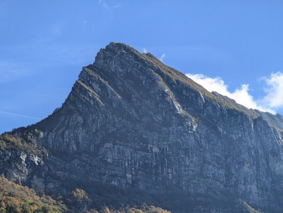 The triangular peak of a limestone mountain rises against a blue sky, above trees whose leaves are yellowing with the autumn changes.