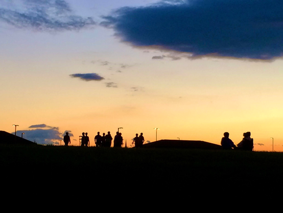 The photograph shows a scenic sunset with a sky transitioning from orange to blue, scattered with a few clouds. In the foreground, there is a silhouette of a hill with multiple people standing or sitting, creating a peaceful, contemplative mood. The people appear as dark shapes against the colorful sky, and a few small streetlights or lampposts are also visible in the distance. The overall feel is calm and serene, capturing a moment of group observation or relaxation at twilight.