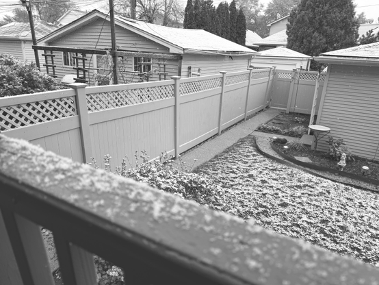 A backyard scene showing a wooden fence, some garden landscaping with a small table and plants, and a dusting of snow on the ground and surfaces in a residential neighborhood. The image is in black and white.