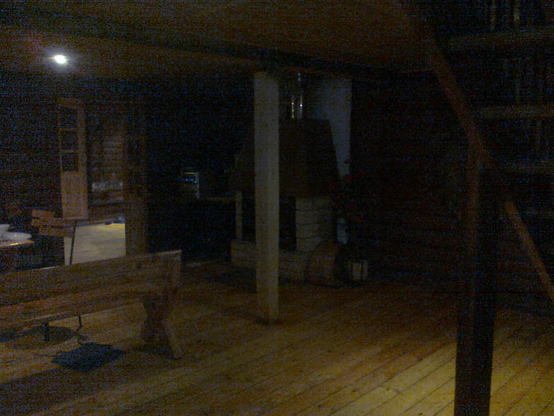 Poor quality photo of a wooden cabin, some stairs visible in the right upper side, a table on the left side with long chairs, a single light fixture over it, further in the dark is a decently sized fireplace and possibly a kitchen further.