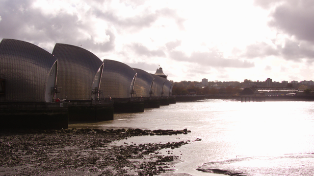 A photograph of the Thames Barrier, taken by me. The low november suy shines off the Thames to the right. The Pontoons of the Thames Barrier stretch away tro the right. There is an errie orange glow on some of the sliver pontoons, a reflection from the sun and the water. 