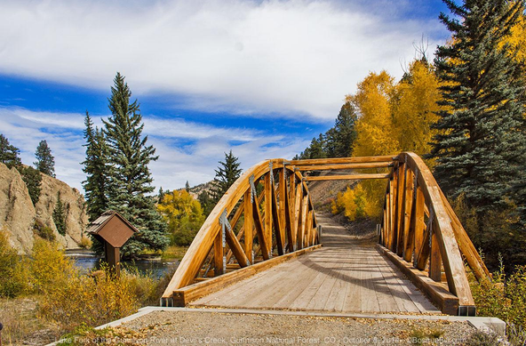 A small wooden arch-truss bridge, only a single lane wide crosses a mountain river connecting a gravel road. Blue spruce and golden aspen can be seen beyond. Low rock cliffs on the left. Cloudy sky with stripes of clear blue.
©BosqueBill.com