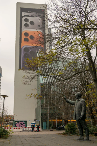 Sculpture/monument of a worker from GDR times, next to a big building with huge iPhone 17 Pro ad on it