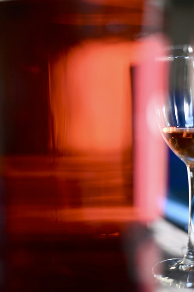 A close-up view of a red-orange bottle glowing with light, its translucent surface filling most of the frame, with a wine glass faintly visible behind it at the edge.