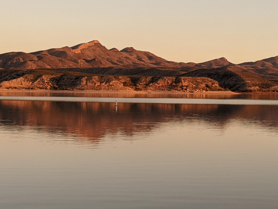 Glowing reddish orange mountains at golden hour reflect onto the shimmering lake water in front of them 