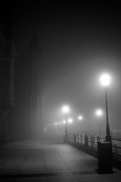 Nightly Motława river bank promenade in fog. Rows of streelamps spread their light in heavy fog, a hint of a tower can be seen on the left. No people are walking by.