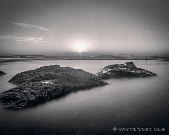 Split toned black and white view of a tidal bathing pool at dawn. The sun is centrally on the horizon, just rising above some cloud into a clear sky. There are two smooth rocky islets in the pool.