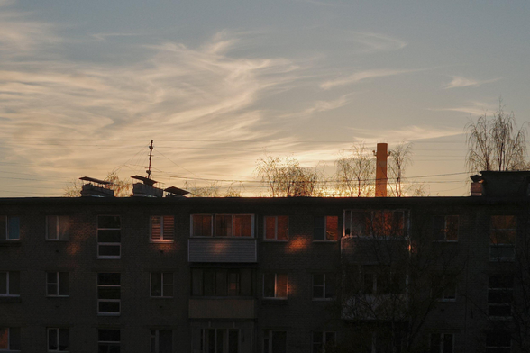 The upper floors of a Soviet-built multi-storey apartment building (Khrushchev buildings) are in the shade. The reflections of the setting sun are visible on the building. The sunset sky is visible above the building. An industrial chimney is visible in the background.