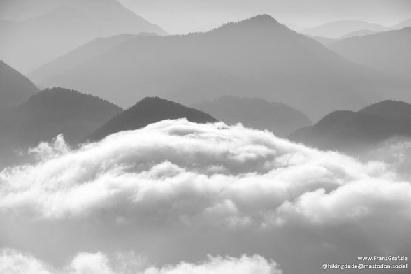 A sea of soft, billowing clouds stretches out beneath a dramatic mountain landscape, their fluffy white peaks gently undulating like waves. The mountains rise majestically from the cloud cover, their rugged silhouettes softened by the misty atmosphere. Layer upon layer of forested ridges and peaks fade into the distance, creating a sense of depth and scale that feels almost infinite.

The scene is serene and ethereal, with the clouds obscuring the lower slopes and adding an air of mystery to the landscape. The light is diffused, casting a gentle glow over the mountains and enhancing the tranquil, dreamlike quality of the moment. The contrast between the solid, dark shapes of the mountains and the soft, flowing clouds creates a striking visual balance, evoking a sense of calm and contemplation.

This breathtaking view captures the quiet grandeur of nature, where the earth and sky come together in a harmonious dance of light, shadow, and mist. It’s a moment that feels timeless, inviting reflection and a deep appreciation for the beauty of the natural world.