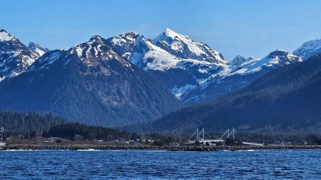 View of Sitka taken from a boat in Sitka Sound. The O'Connell, which connects Japonski and Baranof Islands in is the lower right, just above the water line. You can barely make out buildings along the shoreline, but huge, jagged mountains rise precipitously  in the shot, and the peaks are snow covered. The sky is cloudless and bright blue, giving the whole pic a blue cast when you add in the water. 