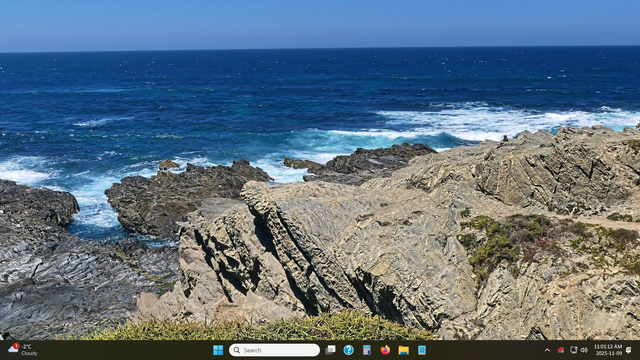 A desktop screenshot showcasing a scenic coastal landscape. The image features a clear blue sky over a vast, deep blue ocean, with waves crashing onto a rocky coastline. The foreground is dominated by rugged, layered rock formations covered in sparse vegetation. At the bottom of the screen is the Windows taskbar.
