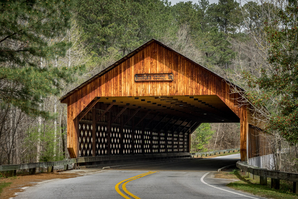 An eye-level, straight-on view of the entrance to a wooden covered bridge. The bridge is stained a warm, rich brown and has a gabled roof. A wooden sign above the dark, rectangular entrance reads "Rockdale County, Ga". The bridge's long sides, visible in perspective, feature a diamond-shaped lattice framework. A two-lane asphalt road with a double yellow line curves from the bottom of the frame and leads directly into the bridge, which is surrounded by a dense forest of tall pines and bare deciduous trees.