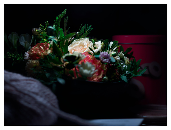 En liggande bild på en låg blomsterbukett som har det enda ljuset från en bordlampa uppifrån. Framför ligger ett lila garnnystan och bortanför en röd kakburk, båda i mörker och lite ur fokus.