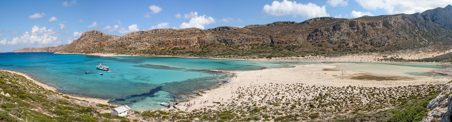 A panoramic view of a coastal landscape featuring turquoise waters, sandy beaches, and rocky hills. Several boats are anchored in the water, and a few people can be seen on the beach and surrounding areas. The sky is partly cloudy.
