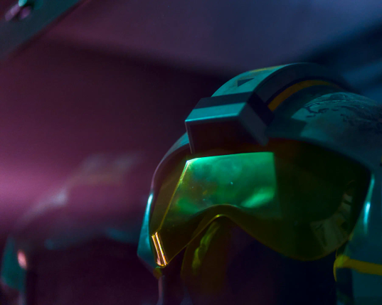 In einem dunklen Raum liegen zwei Helme der Rebellen aus der Filmreihe Star Wars auf einem Regal. Von links fällt magentafarbenes Licht auf die Szene, das Visier des vorderen Helmes leuchtet im Licht grün-metallisch auf. 
.
In a dark room, two helmets worn by rebels from the Star Wars film series lie on a shelf. Magenta light falls on the scene from the left, illuminating the visor of the front helmet in a metallic green glow. 
.