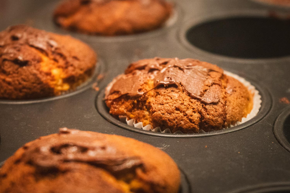 Auf dem Bild liegen frisch gebackene Muffins noch im Backblech. Die Oberfläche ist goldbraun und leicht rissig, einige Muffins sind zusätzlich mit weicher Schokoglasur bestrichen. Das warme Licht betont die Textur des Teigs und vermittelt eine gemütliche, einladende Stimmung – als wären die Muffins gerade erst aus dem Ofen gekommen und bereit zum Genießen.