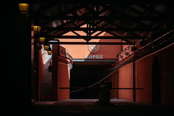 Parking entrance of a port house: we see an orange wall with a signage: "KOPKE estd 1638" and a corridor that pursue in the shadow under it. Outside on the left, a man is sitted on a stairway.