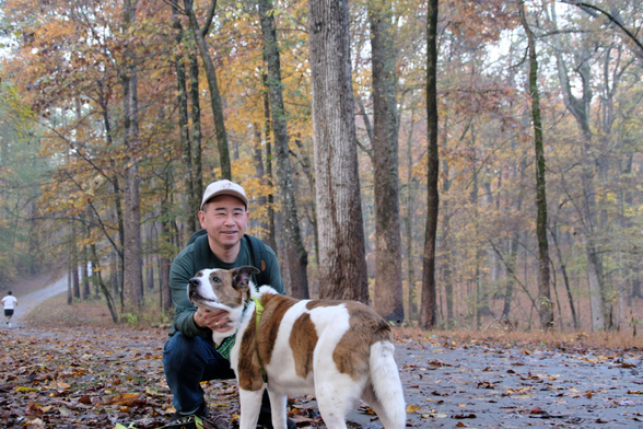 A man with a white cap squatting as he held his white and brown dog on a trail in the woods of a park. A running man is seen at the left edge of the photo