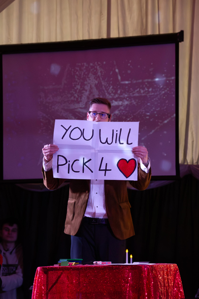 Magician on stage holding a sign saying you will pick 4 of hearts.