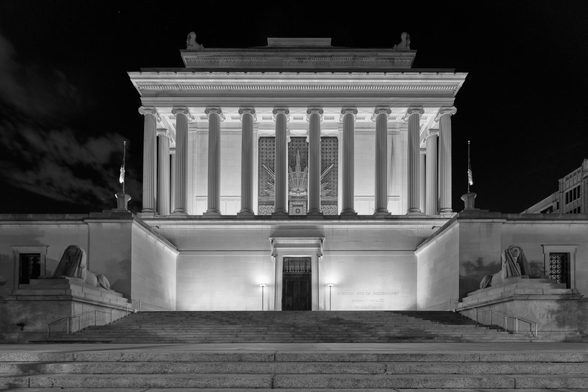 A large Masonic temple at night, with Ionic columns, ornate features, flanked by human lions statuary.