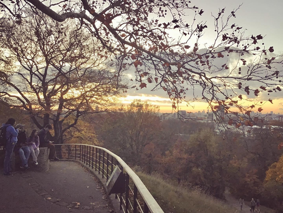 A landscape view across central London commercial skyline, from Greenwich Prime Meridian at dusk, looking across autumn colours. Young people sit and stand and gaze beyond a physical barrier Northwards along an invisible timeline.