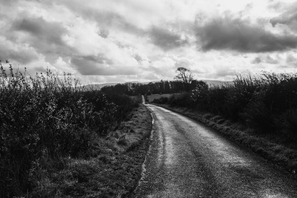 A black and white photograph. A winding road stretches through a landscape with grassy verges under a cloudy sky. 