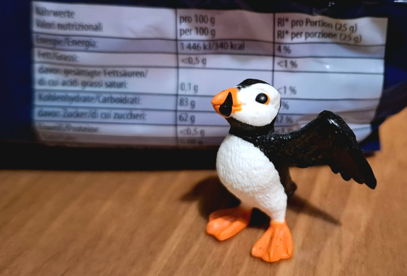 Eine kleine Schleichfigur (Papageientaucher mit ausgestreckten Flügeln) steht auf einer hölzernen Fläche. Dahinter ist eine Plastikpackung, davon aber nur die Nährwerttabelle, zu sehen. U.a. steht beim Wert der Kohlenhydrate 83g pro 100g.