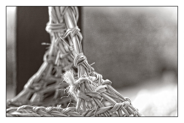 A close-up of woven straw, showcasing intricate knots and textures. The image is in black and white, highlighting the details of the craftsmanship. A blurred background emphasizes the focus on the woven material.