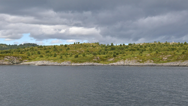 A photo of a rocky shoreline leading up a low green hill. The sky is filled with grey clouds with a little bit of blue sky exposed.