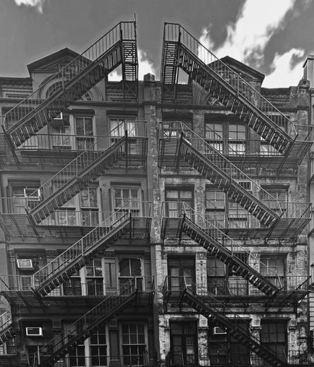 A black and white photo of a building facade featuring multiple fire escapes. Four fire escapes on each side of the photo are angling up towards the middle. So, it almost looks like the fire escapes are making four arrows pointing up on the front of the building. The architectural details includes various windows and a mix of brick and plaster textures. The sky above has some clouds. The dark black and white shading makes the building look especially old 