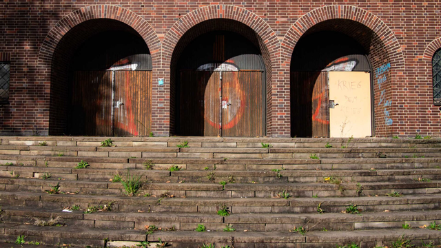 Farbfoto des Portals einer ungenutzen Kirche. Drei Rundbögen aus Backstein überspannen drei Türen, die mit Graffiti verunstaltet sind. Aus den Stufen vor dem Portal wächst Unkraut.
