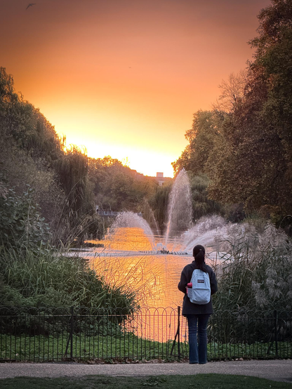 A young female tourist looks at the Swire Fountain in St James's Park under a glowing sunset.