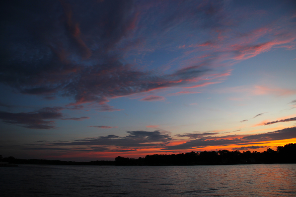 Sunset photo of a dark grey and pink cloud over a sunset and a lake. The cloud looks a bit like a dragon with wings flying to the right.