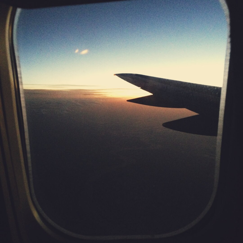 A vertical shot from an airplane window, showing the dark silhouette of the plane's wing tip against a hazy sunset. The sun is a bright spot on the horizon, casting a pale yellow glow that fades into a grainy, deep blue sky at the top of the frame. Below, the ground is obscured by a dark, shadowy expanse of clouds.