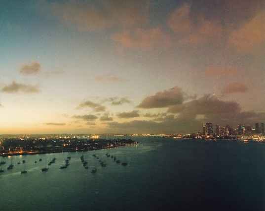 A high-angle, wide shot of a bay at dusk, overlooking a city skyline. In the foreground, dozens of small boats are anchored in the dark, teal-colored water. To the left, a spit of land is dotted with bright city lights. To the right, the illuminated skyline of a downtown area sits on the horizon under a soft, grainy sky with scattered, pink-tinged clouds.