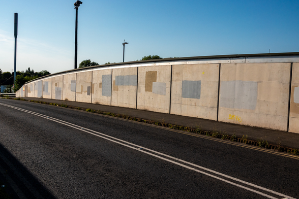 A long road bridge has a tall concrete barrier, in many places along it there are roughly painted rectangles in different shades of faded grey and brown - some overlapping. One has a heart faintly painted over it. Behind, there is a bright blue sky.