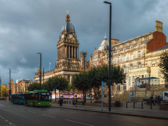 Leeds Town Hall's distinctive Italianate clock tower dominates the centre of this urban scene, its golden-hued stone and ornate cupola crowned with a gilt finial rising majestically against a brooding slate-blue sky mottled with cloud. The magnificent Victorian edifice, constructed from finely dressed ashlar in warm ochre tones, displays the classical architectural vocabulary of its era: fluted columns, decorative stonework, and pitched roof elements adorned with chimney stacks in contrasting red brick. To its right stands an adjacent period building of similar honey-coloured stone with rounded Romanesque-style window arcading, presently undergoing renovation with metal scaffolding visible across its facade. A modern public square fronts these heritage structures, animated with urban life: a green and blue articulated bus moves through the paved forecourt, pedestrians congregate amongst contemporary street furniture and lamp standards, whilst mature deciduous trees provide patches of autumnal golden foliage that complement the warm stonework. The composition captures a perfect marriage of civic Victorian grandeur and contemporary urban vitality, the architectural heritage anchored firmly within the rhythms of modern city life.
