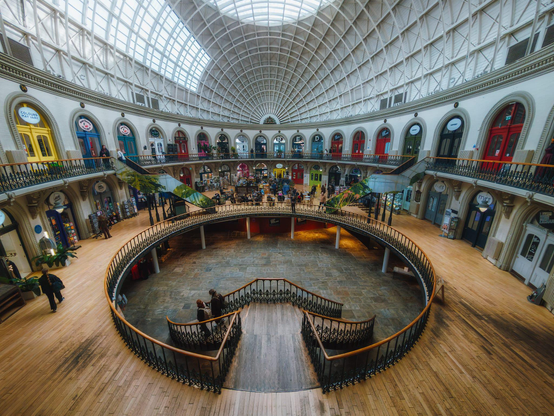 The Corn Exchange in Leeds displays its remarkable Victorian circular arcade architecture beneath an expansive glazed barrel-vaulted ceiling of cast iron and glass that floods the interior with natural light, creating a luminous cathedral-like atmosphere. The building's distinctive ringed gallery levels feature a succession of brightly painted arched shop frontages in jewel tones—yellows, blues, teals, reds, and greens—each surmounted by ornamental pediments and separated by decorative cornicing that runs continuously around the cylindrical space. Wrought-iron railings with burnished brass rails delineate each level, their serpentine curves echoing the building's circular geometry, whilst the warm honey-toned wooden floors of the ground level contrast beautifully with the darker slate-grey patterned flooring of the central sunken floor. A sweeping curved double staircase with elegant brass handrails descends into this central arena, where visitors congregate, creating a focal point beneath the soaring geometrical tracery of the iron roof structure. Potted plants and contemporary retail signage animate the galleries above, where shoppers browse the specialist independent retailers that populate this Grade II* listed building, transforming this former mercantile space into a vibrant contemporary shopping destination whilst preserving its exceptional architectural integrity.
