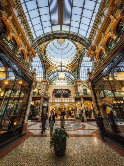 County Arcade in Leeds presents a sumptuous Victorian arcade of exceptional architectural refinement, its soaring barrel-vaulted glass roof supported by ornamental cast-iron ribs flooding the interior with soft, diffused natural light that illuminates the richly decorated terracotta and cream-coloured faience tilework adorning the vaulted ceiling. The corridor stretches away in symmetrical perspective, flanked on both sides by elegant shop frontages with mahogany-framed glazing and gilt detailing, their display windows glowing warmly beneath brass light fittings that cast pools of amber illumination across the tessellated terracotta and cream geometric floor patterning below. Ornamental green and gilt decorative panels and scrollwork embellish the walls between shopfronts, creating a rhythm of pattern and refinement typical of late-Victorian arcade design, whilst cream-coloured classical pilasters with decorative capitals provide structural and visual emphasis to the elevation. The arcade is animated with shoppers moving between the luxury retailers—visible signage includes Reiss amongst other boutique establishments—their silhouettes contrasting against the honey-toned architectural backdrop, whilst a potted ivy plant marks the central axis of the perspective, anchoring the composition with a touch of living green against the sumptuous architectural theatre surrounding it.
