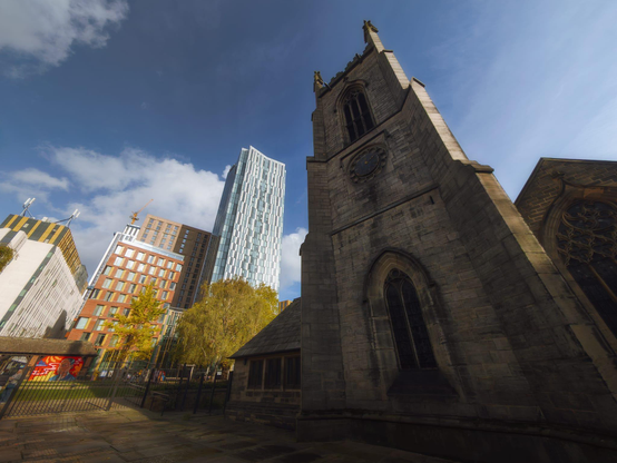 St. John the Evangelist's Church in Leeds rises majestically in coursed gritstone Gothic Revival architecture, its steeply pitched roof and soaring pinnacled tower dominating the composition against a bright cerulean sky streaked with white cloud, the tower crowned with gilt finials and pierced by elegant lancet windows characteristic of the Perpendicular style. The church's robust masonry, rendered in grey-buff stone with darker weathered patches, displays the typical vocabulary of Victorian ecclesiastical design—pointed arch windows with geometric tracery, buttresses articulating the elevation, and decorative carved stonework—whilst the substantial tower features a prominent circular clock face set within the masonry. To the immediate left stands a contemporary iron railing enclosing the churchyard, beyond which modern residential and commercial architecture rises in stark contrast: sleek glass-clad high-rise towers in pale blue and cream, warm terracotta-brick mid-rise apartment blocks, and contemporary mixed-use developments with pitched roofs echoing the church's silhouette. Deciduous trees with autumn-golden foliage soften the urban streetscape, their warm tones complementing the gritstone church walls, creating a compelling dialogue between Leeds' nineteenth-century ecclesiastical heritage and its rapidly evolving contemporary skyline, the church standing as a steadfast sentinel amidst the city's ongoing architectural transformation.