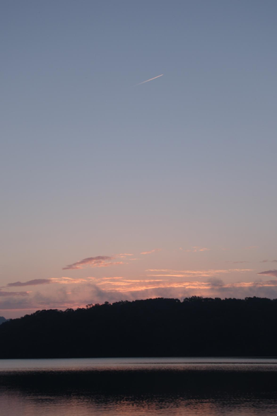 A lakeview at dawn as the sun tries to show above the woods turning the clouds pink. A short contrail left by an airplane is seen near the top of the photo.