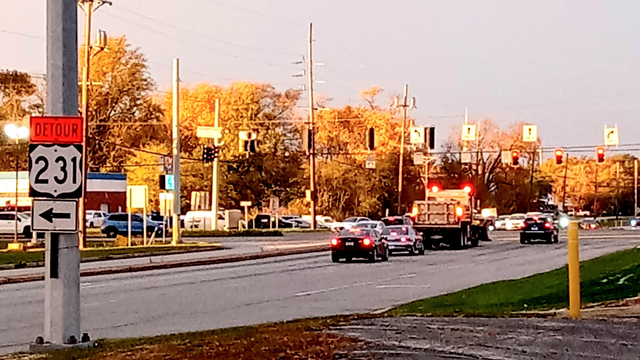 A salt truck with a snow plow prepares the streets for a lake effect snow storm forecasters predict will bring 8" to 12" of snow starting tonight. The scene is at Taft and Lincoln Hwy (US 30) in Merrillville. Cars are stopped at the traffic light. A detour sign for US 231 points left (west). Trees with golden fall colors are in the background. Commercial businesses are on the corners. 