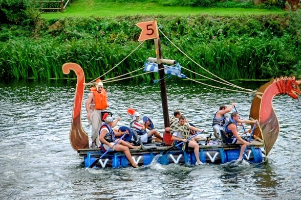 A homemade, Viking-themed raft built from blue barrels, decorated with a dragon head and number '5' flag, being paddled down the River Wye by several enthusiastic people in life vests. One person at the rear is dressed in a Viking helmet, adding to the silly and festive atmosphere.