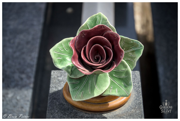 A focused, overhead close up of a single ceramic rose serving as a grave decoration.

The bloom features dark, rich, maroon-coloured petals spiralling inward, set against five vibrant green leaves with etched veins.

The ceramic piece rests on a polished wooden base atop dark grey granite stonework. The background shows blurred parallel lines of granite. Signed Bron Peirce.
