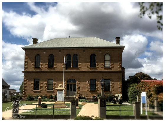A two story, yellowish brown sandstone building with a corrugated iron hip roof, the Oatlands Council Chambers, stands under a dramatic, cloudy sky.

A flagpole is centred in front of the building, which is framed by a low green fence and a paved walkway leading to the entrance. The building has symmetrical arched windows.