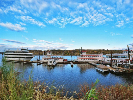 One of a series of scenes from the Stillwater, Minn., riverfront area, featuring a bridge crossing the Mississippi River, riverboats docked at a marina, and rusty chains connecting concrete pillars along the shoreline’s walkway.
