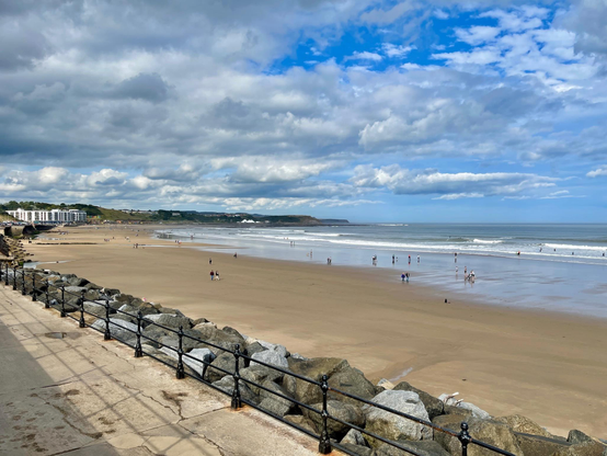 A wide sandy beach stretches along the coastline, with gentle waves reaching the shore under a partly cloudy sky. People are scattered across the wet and dry sand, some walking near the water and others standing in small groups, while a few venture into the shallow surf. A concrete promenade with black railings runs parallel to the beach, bordered by large rocks serving as a sea defense. In the distance, low cliffs and rolling hills frame the horizon, and a cluster of white buildings sits near the shoreline. The tide appears to be out, exposing a broad expanse of sand, and the scene conveys a calm, leisurely atmosphere.