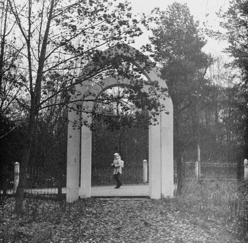 In the black-and-white photo, a white archway with a Star of David is the entrance to an old Jewish cemetery (seen from the cemetery side). A fence is adjacent to the archway. A woman is walking past the cemetery, in the center of the archway. Trees can be seen on the sides and in the distance. Fallen leaves are on the ground.