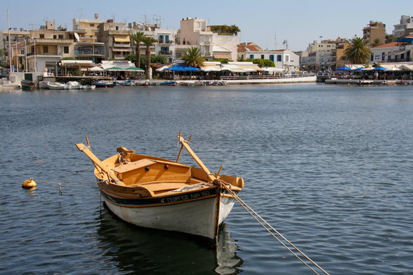 A small wooden boat is docked in a calm harbor with buildings and palm trees in the background. The scene features clear blue skies and water.